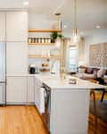 Neutral colored cabinets with brass hardware in open kitchen with open shelving and two large bulb pendant lights over countertop.