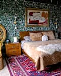 Bedroom with green floral wallpaper and wicker chair in corner beside bed. Bed is made with tan velvet blanket and animal print linens. Vintage wooden dresser across from bed.