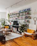 Cozy living room with bookshelves, plants, a gray sofa, wooden floors, and a vintage "Ralph's Ice Cream" sign.