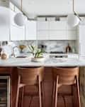 Kitchen with marble bar/counter/backsplash, curve-backed wooden stools, white cabinets, fridge, white globe pendant lights