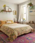 Plant filled bedroom with neatly made bed topped with pale yellow quilt. Colorful pink rug lines the floor.
