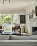 Living room with white walls, gray painted brick fireplace, gray sofa, green front door, large windows, 2 wood white cushioned armchairs, wood floors