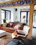 Living room with overhead wood beam decorated by blue and white plates, rust leather sofa, white cube coffee table, leather armchair with throw pillows, hanging plants, large windows, white walls