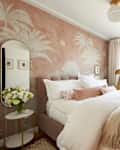 Bedroom with velvet quilted headboard and bed frame, white linens, blush velvet pillow, oval mirror on wall over small white table with vase of white flowers. Wall has palm tree themed wallpaper in blush and cream. Large window with off white curtains
