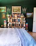 Bedroom with neatly made bed and lots of books on open shelves surrounded by landscape paintings.