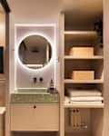 Modern bathroom with round mirror, green vanity, wall art, and open shelving with towels and wooden boxes.