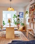 White dining room with large wood table and rattan backed chairs on sisal rug. Lots of plants around