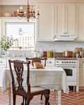 A white dining room table for two near a kitchen with all white cabinets and appliances on a tile floor.