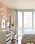 Bright bedroom featuring a floral accent wall, a bed with patterned bedding, a desk with mirrors, and large windows.