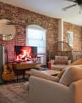Cozy living room with exposed brick walls, a TV displaying a fireplace, beige sofa, wicker chair, and a guitar.