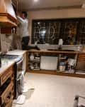 Cluttered kitchen with wooden cabinets, a man fixing a cabinet, and various items on the countertop.