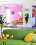 Colorful living room with green sofa, yellow pillows, tulips, pink walls, and a retro dining area with a round table and chairs.