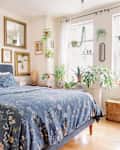 Cozy bedroom featuring a blue floral bedspread, framed art on the walls, and various indoor plants by the windows.