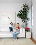 A woman and a girl play on a white sectional sofa in a bright living room with a large abstract painting and a potted plant.