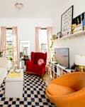 Cozy living room with a white sofa, red armchair, orange accent chair, and a checkered rug. Shelves display books and decor.