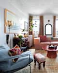 Cozy living room with a pink sectional sofa, colorful pillows, a round coffee table, and plants by the windows.