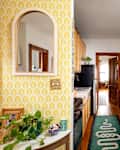 Narrow kitchen hallway with yellow patterned wallpaper, wooden cabinets, green tile backsplash, and plants on shelves.
