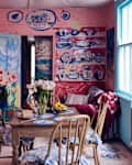A colorful dining area with pink walls adorned with abstract art, a wooden table set with flowers, fruit, and cake.