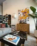 Marble coffee table and gray sofa in front of shelving unit across from large floor mirror, wall sconce, art, bar cabinet, and potted tree.