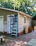 A green garage with peeling paint, a ladder against the wall, and wooden furniture pieces nearby.