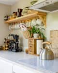 Bright kitchen corner with a wooden shelf holding mugs, bowls, and a kettle, featuring plants and a coffee maker.