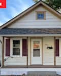 A small, white house with a gabled roof, burgundy shutters, and a front porch featuring lattice skirting.
