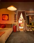 Cozy bedroom with a bed, colorful pillows, a red nightstand, and a green sofa, illuminated by warm lighting.