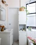 Bright bathroom with a white sink, towel, and plants in a cabinet, featuring a window and floral decor in the bathtub.