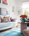 Bright living room featuring a light gray sofa with colorful pillows, a round coffee table, and vibrant artwork on the walls.