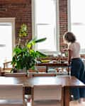 A woman with curly hair prepares food in a bright, rustic dining area with brick walls, plants, and a wooden table.