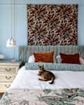 Cozy bedroom featuring a striped headboard, decorative pillows, a cat on the bed, and a floral wall hanging.