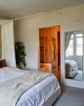 Cozy bedroom featuring a large bed with white linens, decorative pillows, and a plant, with an adjoining bathroom visible.