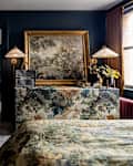 Cozy bedroom featuring a floral bedspread, ornate dresser, and a closet with colorful clothing. A landscape painting and lamp add elegance.