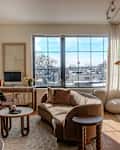 Cozy living room with a curved brown sofa, marble coffee table, desk with a computer, and large windows.
