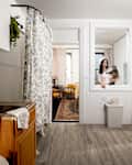 A woman holding a cat stands in a kitchen with white cabinets, a sink, and a floral shower curtain.