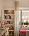 Cozy living room with a bookshelf, large window, pink throw, and a lamp, featuring decorative items and plants.