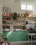 Bright kitchen with green cabinets, stainless steel appliances, wooden shelves, and colorful artwork on the walls.