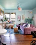 Brightly decorated living room with floral wallpaper, a purple sofa, a round dining table, and colorful artwork.