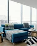 Modern living room with blue sectional sofa, patterned pillows, round coffee table, and striped rug near large windows.