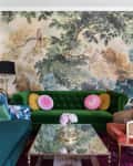 Eclectic living room with green velvet sofa, floral wallpaper, glass coffee table, and colorful cushions.
