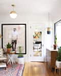 Bright living room with bookshelf, framed art, plants, and a patterned rug. Mid-century chair and wooden dresser by the window.