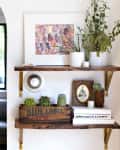 Wooden shelves with potted plants, framed art, books, and a vintage crate in a living room with a gray sofa and colorful cushions.