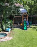 Backyard with a wooden playset, green slide, swings, small pool, and blue chairs surrounded by lush greenery.