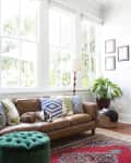 Living room with brown leather sofa, colorful pillows, green ottoman, large windows, and a dog resting on the couch.
