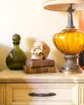 Skull on vintage books beside amber lamp and green glass vase on a wooden dresser.