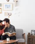 Couple sitting on a gray sofa, woman in red dress, man reading a book, wall with eclectic decor and framed art.
