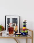 Bar cart with assorted liquor bottles, a framed artwork, colorful mugs, green apples, and a glass decanter.