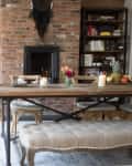 Dining room with brick wall, wooden table, candles, flowers, and a large mirror. Bookshelf and fruit on the table.