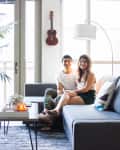 Couple sitting on a gray sectional sofa in a bright living room with a guitar on the wall and a large potted plant.