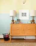 Mid-century wooden sideboard with two lamps, framed art, and a potted plant against a two-tone yellow and blue wall.